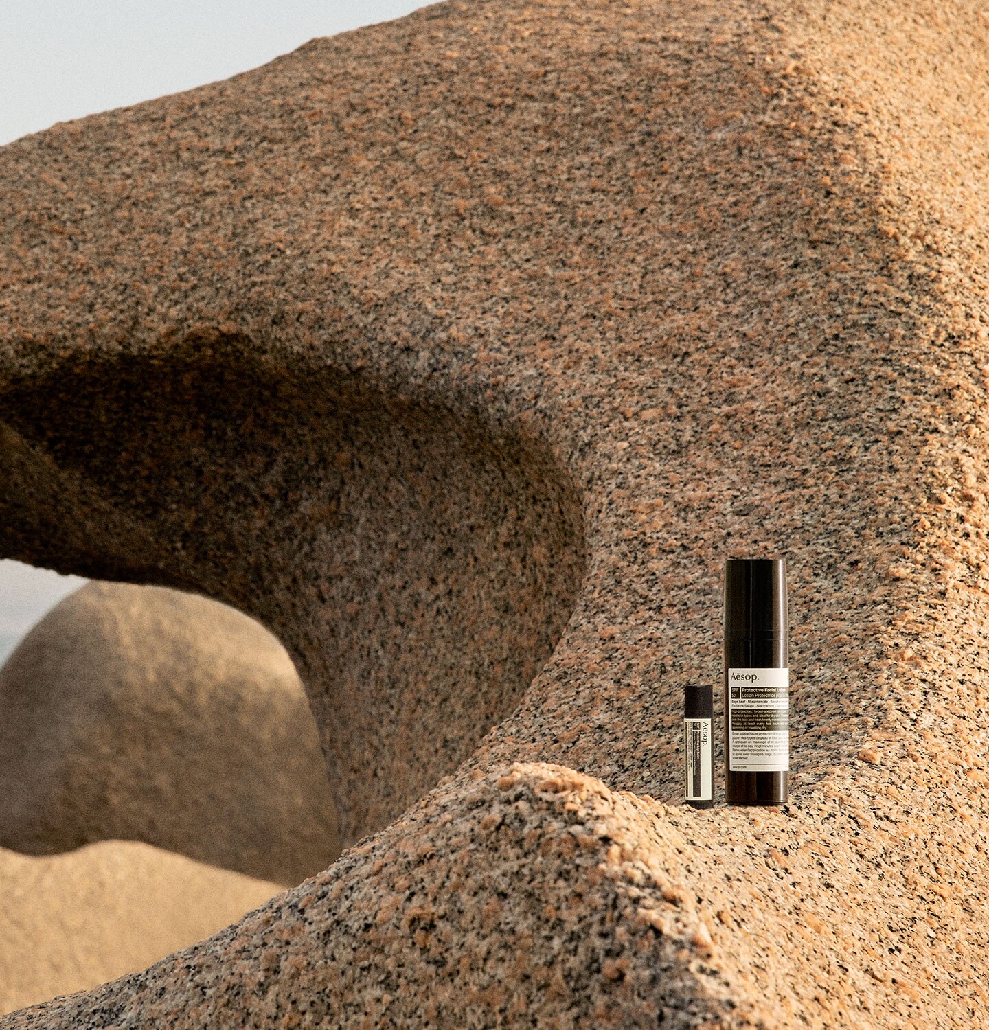 A bottle of Protective Facial Lotion SPF50 and Protective lip balm spf 30 resting on a rocky shoreline.