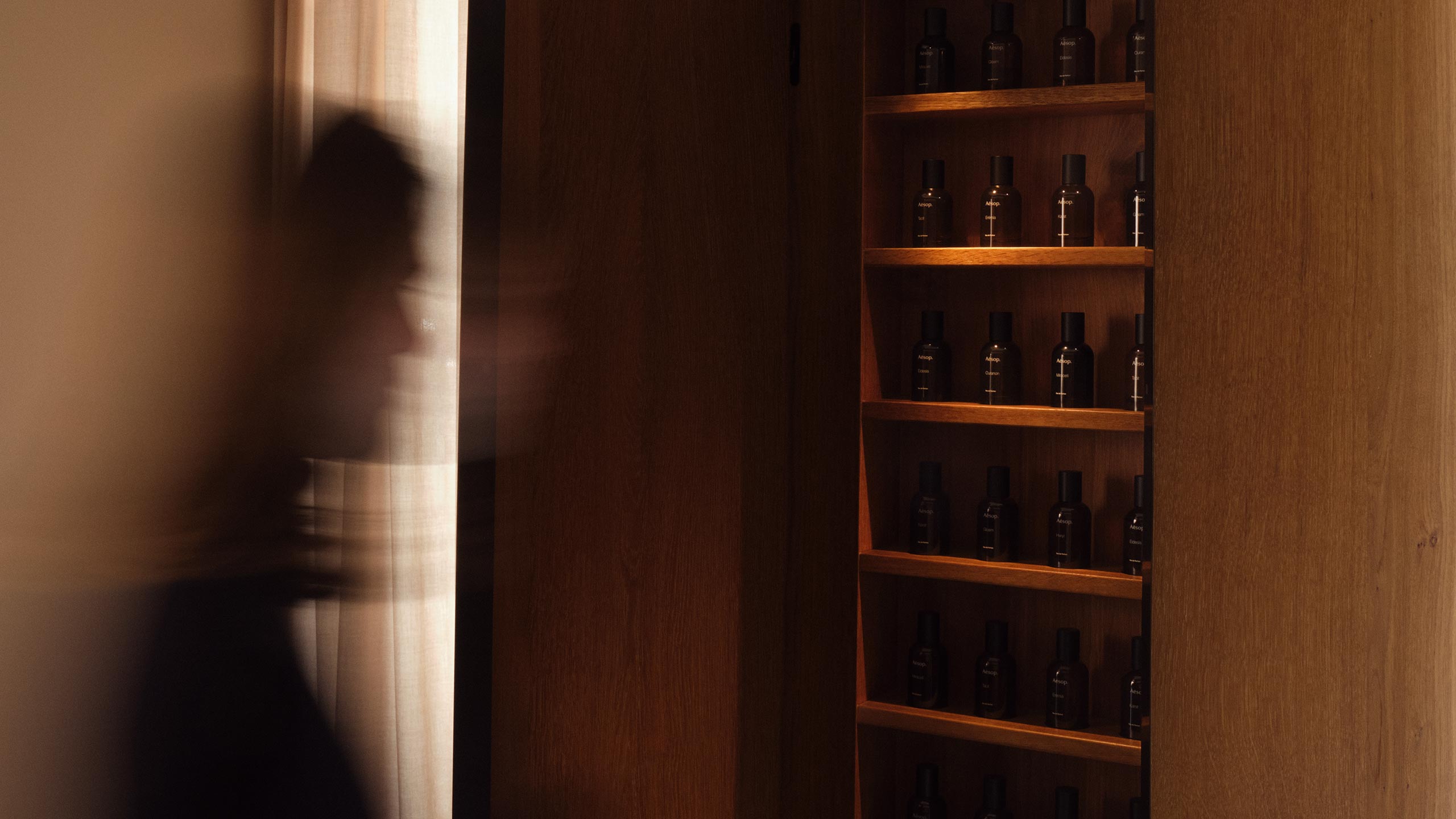 An individual walking past an wooden shelf displaying Aesop fragrances in amber bottle